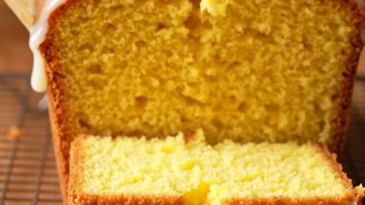 A golden whole lemon blender pound cake with white glaze on a cutting board, one slice cut showing moist crumb.
