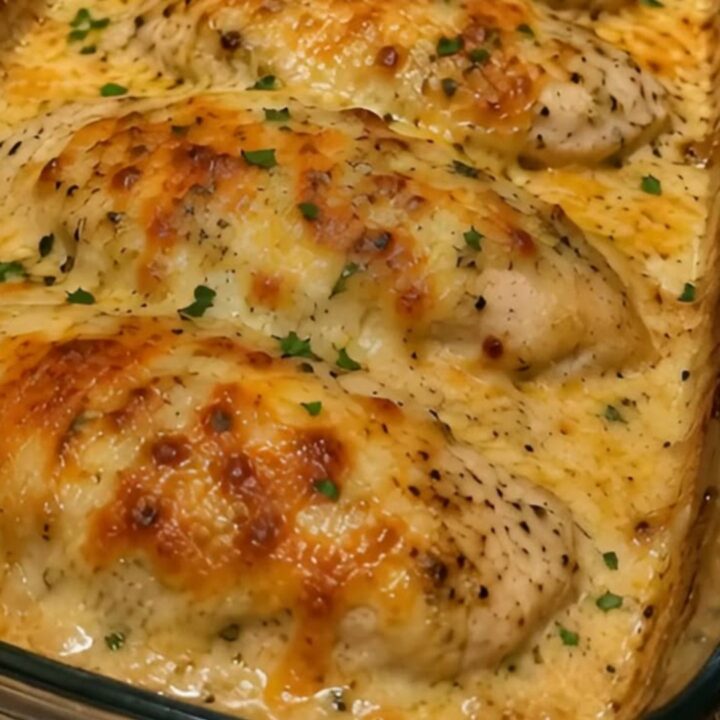 Four golden, cheesy baked chicken breasts in a glass dish on a granite surface.