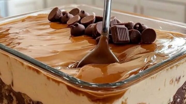 A close-up of a square glass dish filled with layers of cookie crust, whipped cream, caramel, and chocolate.