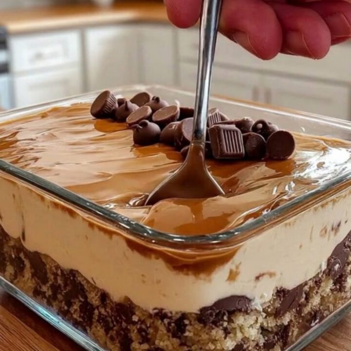 A close-up of a square glass dish filled with layers of cookie crust, whipped cream, caramel, and chocolate.