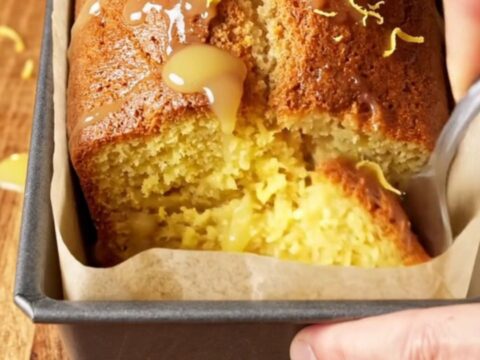 Whole lemon blender loaf cake with white glaze and lemon zest on top, sliced to show moist yellow crumb.