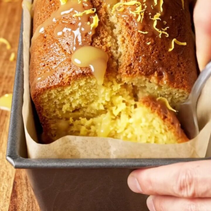 Whole lemon blender loaf cake with white glaze and lemon zest on top, sliced to show moist yellow crumb.