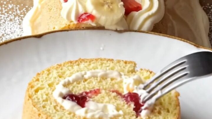 Swiss roll dusted with powdered sugar, topped with cream rosettes and berries, slice showing fruit filling