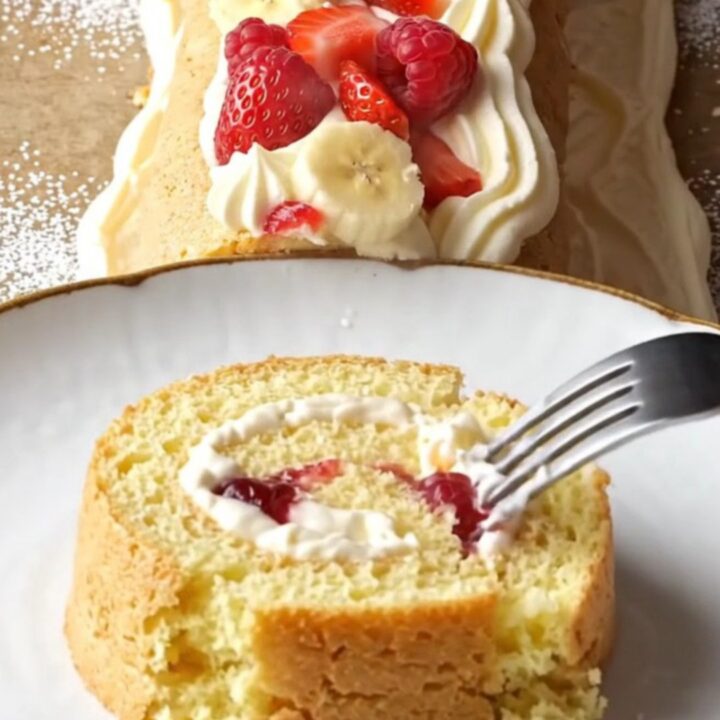 Swiss roll dusted with powdered sugar, topped with cream rosettes and berries, slice showing fruit filling
