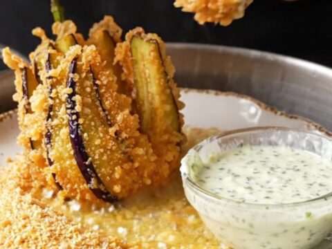 A golden fried eggplant fan on a white plate with a bowl of garlic herb yogurt dip