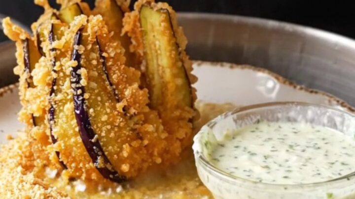 A golden fried eggplant fan on a white plate with a bowl of garlic herb yogurt dip