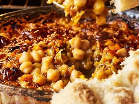 Golden chickpea casserole with roasted peppers and onions in a glass dish, with a piece of flatbread scooping up the mixture.