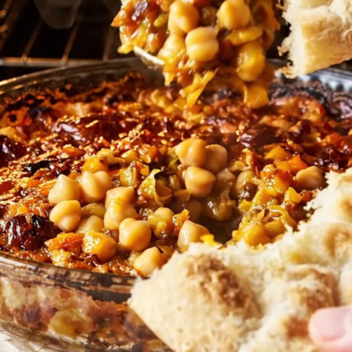 Golden chickpea casserole with roasted peppers and onions in a glass dish, with a piece of flatbread scooping up the mixture.