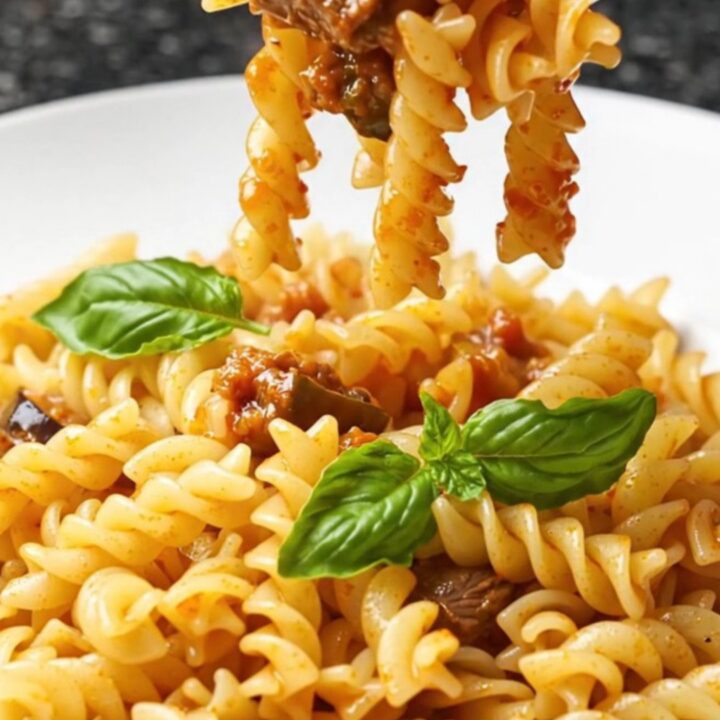 A fork twirls fusilli coated in a rich tomato and eggplant ragù with basil leaves