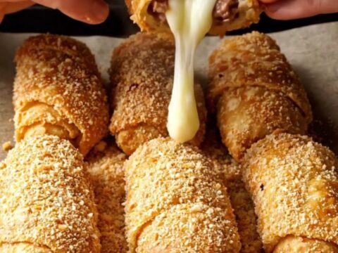 Golden crispy breaded meat börek rolls on a baking sheet, one snapped in half showing melted cheese.
