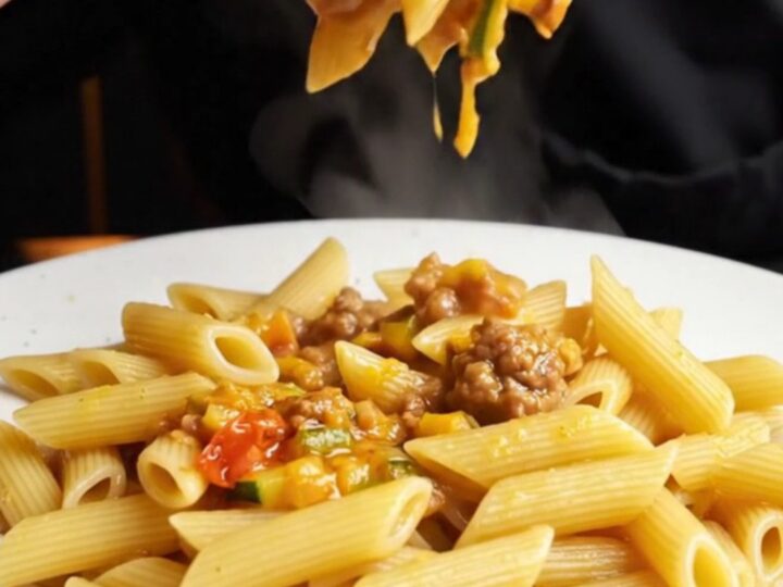 A skillet of penne pasta with chunky tomato-zucchini ragù and beef meatballs, garnished with fresh basil.
