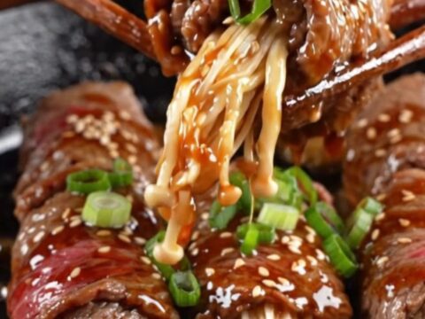Glazed beef-wrapped enoki mushrooms on a plate, garnished with sesame seeds and scallions.