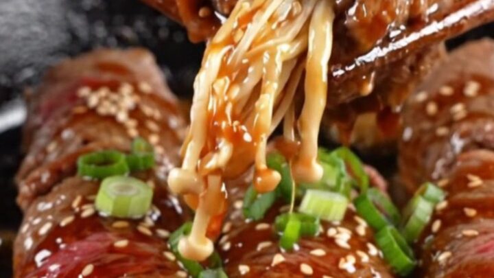 Glazed beef-wrapped enoki mushrooms on a plate, garnished with sesame seeds and scallions.