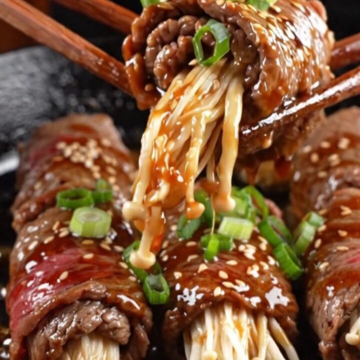 Glazed beef-wrapped enoki mushrooms on a plate, garnished with sesame seeds and scallions.