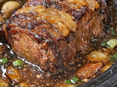 A tender beef pot roast in a black slow cooker, glistening with garlic butter broth and topped with fresh scallions.