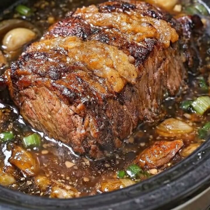 A tender beef pot roast in a black slow cooker, glistening with garlic butter broth and topped with fresh scallions.