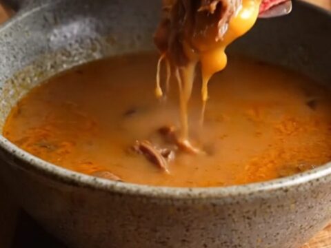 Crispy birria quesadilla on a cutting board, sliced to show melted cheese and shredded beef, with a bowl of consommé