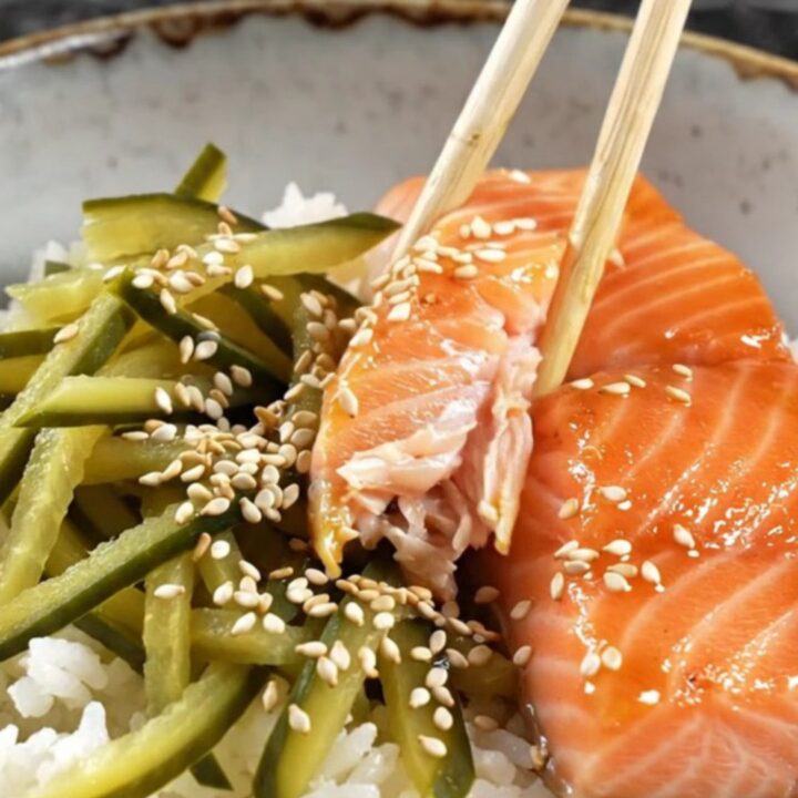 A bowl of teriyaki salmon with glossy glaze, pickled cucumber ribbons, and sesame seeds over rice