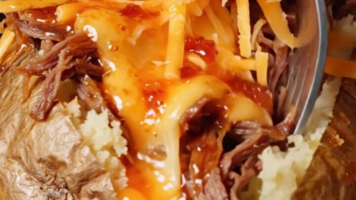 A baked potato split open, loaded with shredded beef and melted cheese, with consommé drizzled over the top.