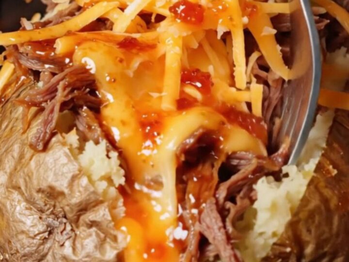 A baked potato split open, loaded with shredded beef and melted cheese, with consommé drizzled over the top.