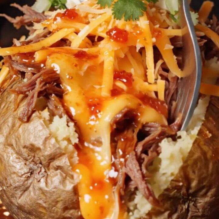 A baked potato split open, loaded with shredded beef and melted cheese, with consommé drizzled over the top.