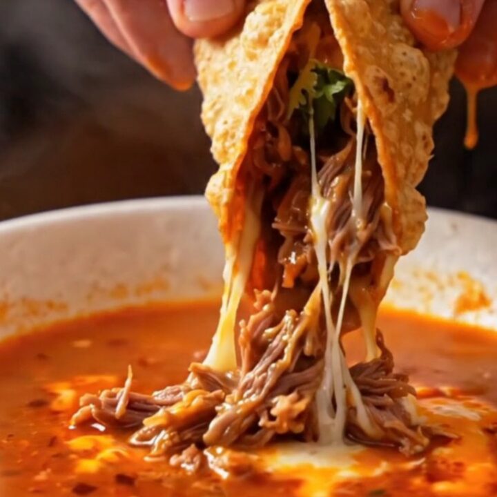 A crispy birria quesataco dipped into a bowl of rich consommé, with melted cheese and beef visible.
