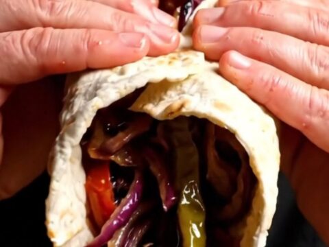 Sizzling Adana kebab on a wide skewer being slid onto warm lavash with charred tomatoes and sumac onions.