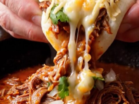 A steamed bao bun stuffed with birria beef and melted cheese, dunked into a bowl of red consommé.