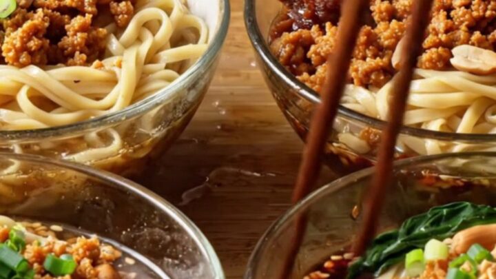 A bowl of Szechuan Dan Dan Noodles topped with crispy pork, crushed peanuts, and green bok choy