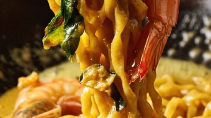 Golden salted egg yolk prawn noodles in a dark bowl, garnished with crispy curry leaves and sliced chilies