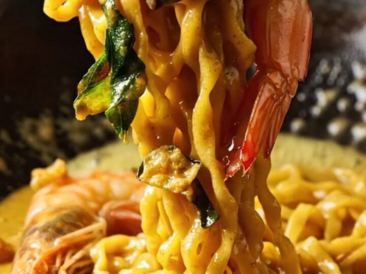 Golden salted egg yolk prawn noodles in a dark bowl, garnished with crispy curry leaves and sliced chilies