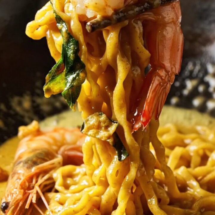 Golden salted egg yolk prawn noodles in a dark bowl, garnished with crispy curry leaves and sliced chilies