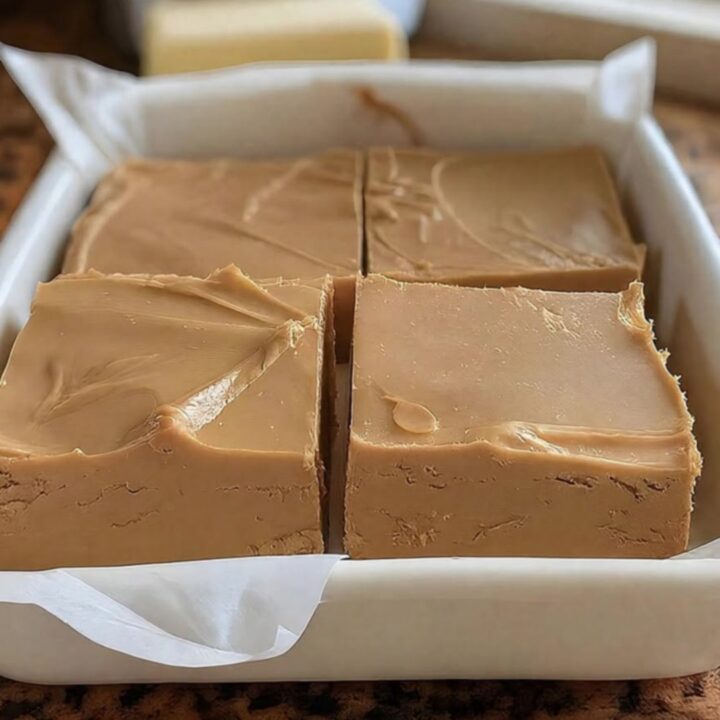 A white ceramic dish filled with thick, perfectly sliced squares of creamy peanut butter fudge near a sunlit window.
