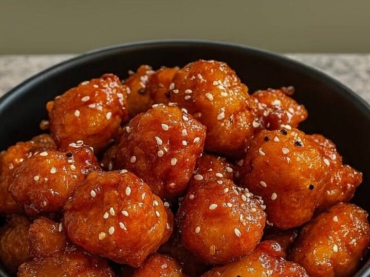A black bowl filled with crispy honey garlic chicken bites topped with sesame seeds on a granite counter by a window.