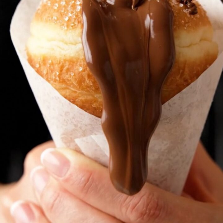 A sugar-coated donut in a paper cone, oozing chocolate hazelnut spread