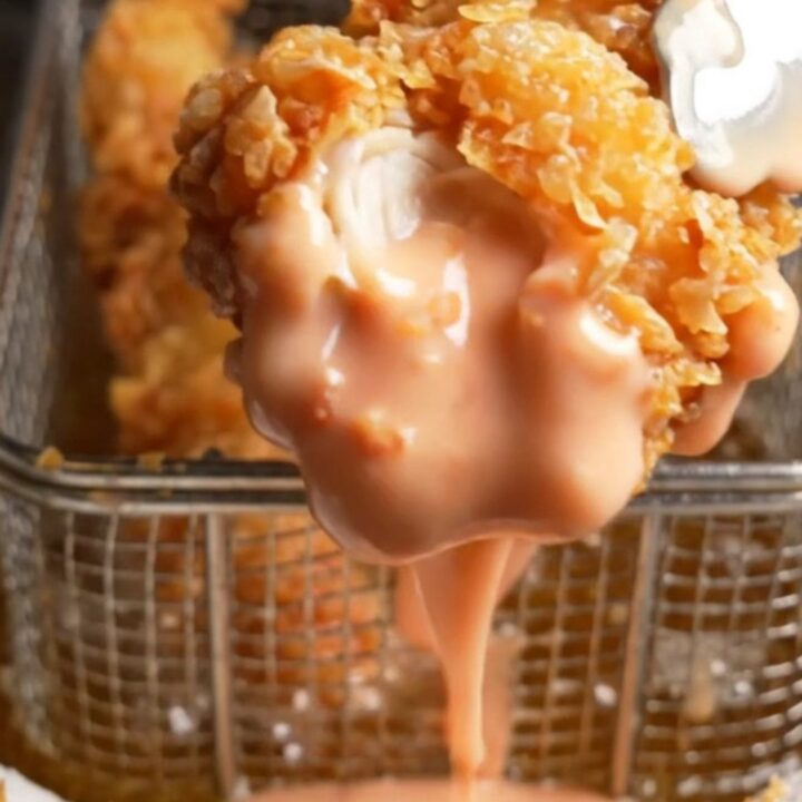 Golden fried chicken fingers stacked on a plate with a small bowl of pink Cane's sauce for dipping.