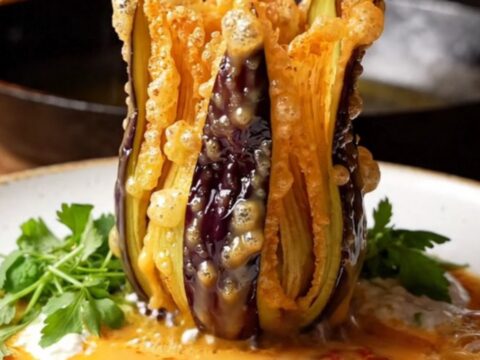 A crispy blooming eggplant fan drizzled with garlic yogurt and sizzling pepper butter, served on a white plate.