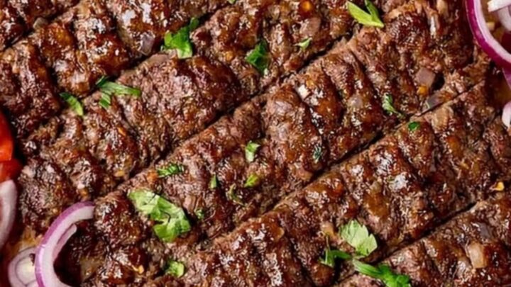 A close-up shot of freshly baked, juicy kofta kebabs on a parchment-lined tray, garnished with fresh parsley and red onion.