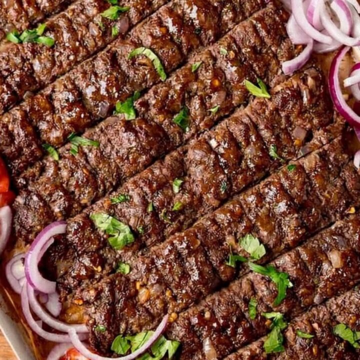 A close-up shot of freshly baked, juicy kofta kebabs on a parchment-lined tray, garnished with fresh parsley and red onion.