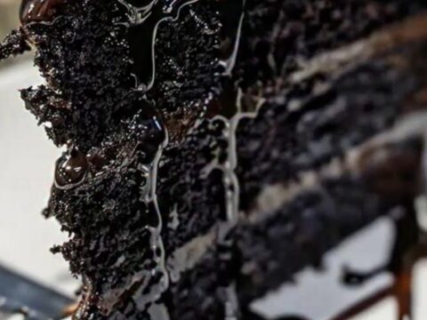 A towering slice of black velvet cake on a white plate with glossy chocolate ganache drips.