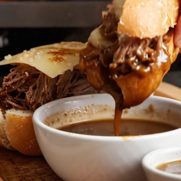 A crusty baguette sandwich filled with shredded short rib and melted Gruyère, being dipped into a bowl of dark au jus.