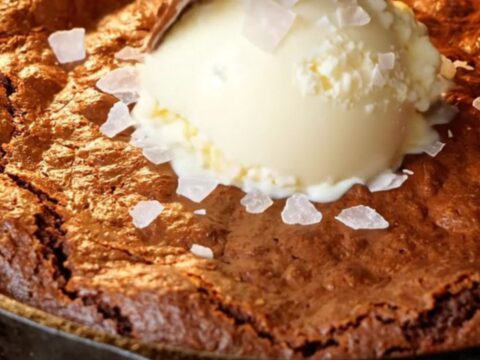 Warm skillet brownie with a crackly top and a spoon pulling up molten fudge, topped with vanilla ice cream and flaky sea salt