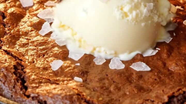 Warm skillet brownie with a crackly top and a spoon pulling up molten fudge, topped with vanilla ice cream and flaky sea salt