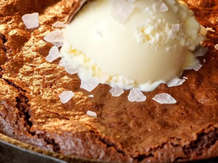 Warm skillet brownie with a crackly top and a spoon pulling up molten fudge, topped with vanilla ice cream and flaky sea salt