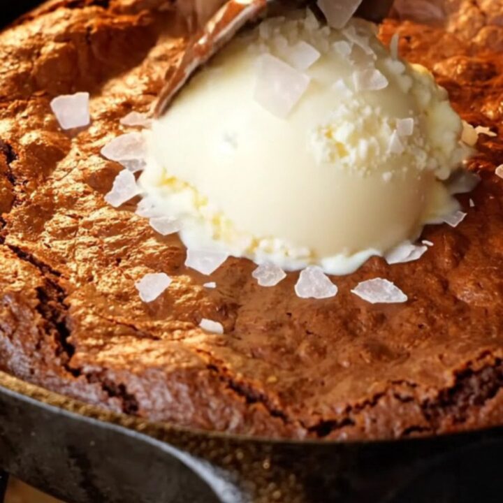 Warm skillet brownie with a crackly top and a spoon pulling up molten fudge, topped with vanilla ice cream and flaky sea salt