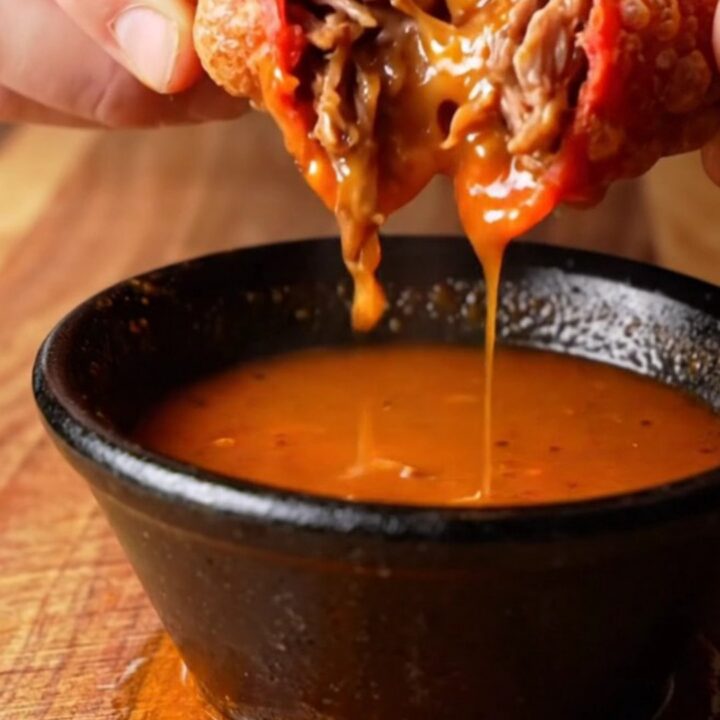 Crispy birria mulitas sliced in half, showing layers of beef and melted cheese, with a bowl of consommé for dipping