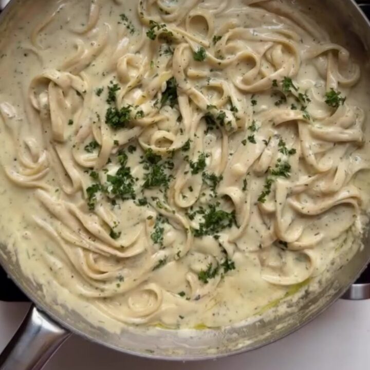 A fork twirling glossy fettuccine Alfredo with a creamy white sauce, garnished with black pepper and parsley.