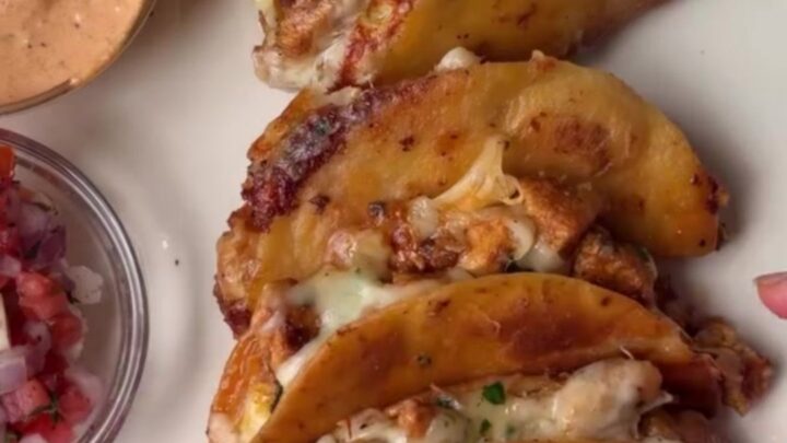 A stack of crispy golden mini tacos with melted cheese and chipotle chicken, served with a bowl of lime crema.