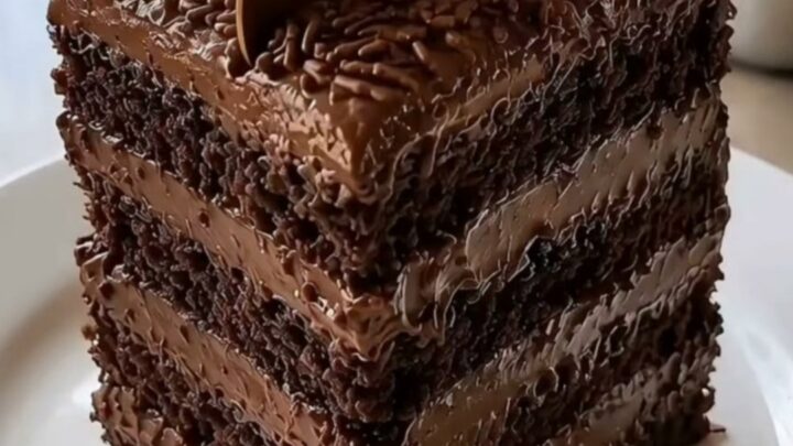 A tall, multi-layer square chocolate cake topped with brigadeiro truffles and a gold spoon on a white plate.