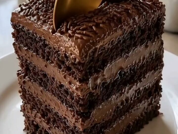 A tall, multi-layer square chocolate cake topped with brigadeiro truffles and a gold spoon on a white plate.